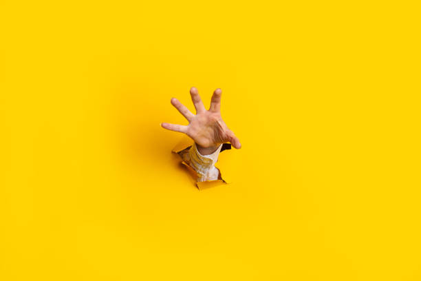 la main de la femme est étendue et saisit quelque chose d’un trou dans le mur sur un fond jaune. bannière - hole punched paper photos et images de collection