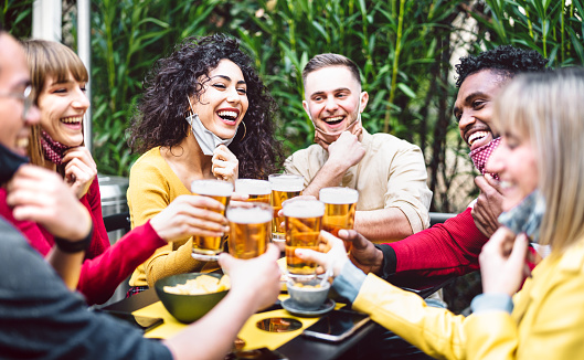 Young people toasting beer wearing open face mask - New normal life style concept with friends having fun together outside at brewery bar garden - Warm filter with focus on woman in yellow clothes