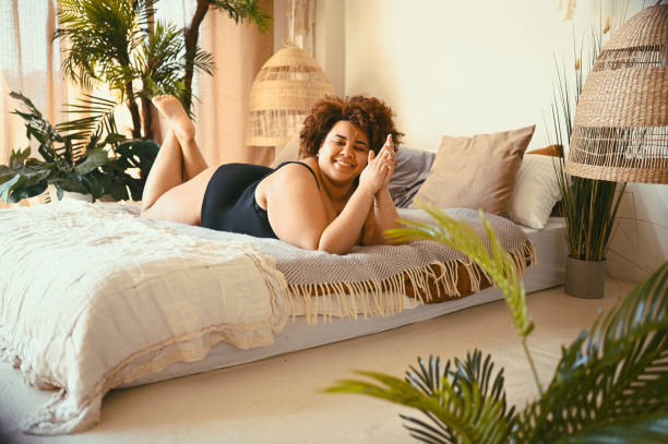 Beautiful curvy oversize African black woman afro hair lying on bed eco friendly bedroom cozy interior design with home plants. Body imperfection, body acceptance, body positive and diversity concept stock photo