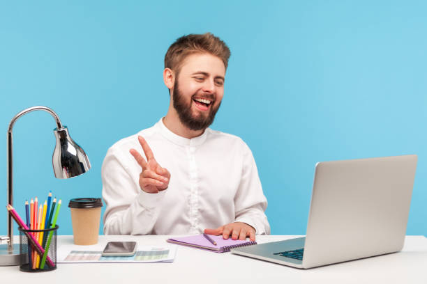 istock Positive bearded man office worker showing v gesture with fingers talking video call, looking at laptop web camera with toothy smile sitting at workplace 1309558784