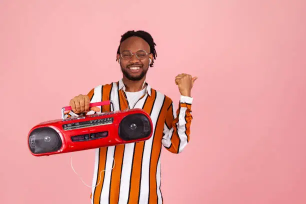 Smiling afro-american man with dreadlocks in striped shirt holding tape recorder listening to music in earphones pointing at freespace for advertisement. Indoor studio shot isolated on pink background