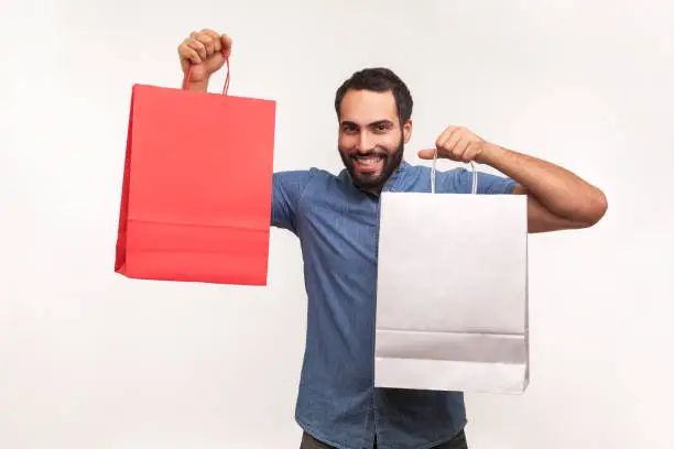 Photo of Satisfied bearded man in blue shirt bragging with paper shopping bags, satisfied with black friday big sale