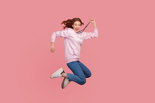 Full length portrait of extremely happy teen girl with curly hair in hoodie jumping for joy or flying, celebrating success, victory, Indoor studio shot isolated on pink background
