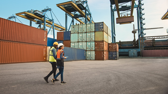 Multiethnic Female Industrial Engineer with Tablet and Black African American Male Supervisor in Hard Hats and Safety Vests Walk in Container Terminal. Colleagues Talk About Logistics Operations.