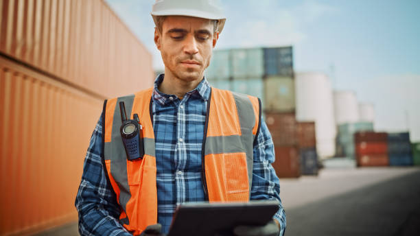 istock Handsome Caucasian Industrial Engineer in White Hard Hat, Orange High-Visibility Vest, Checkered Shirt and Work Gloves Working on Tablet Computer in Shipping Cargo Container Terminal. 1309560053
