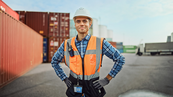Portrait of a Handsome Caucasian Industrial Engineer in White Hard Hat, Orange High-Visibility Vest, Checkered Shirt, Jeans and Work Gloves. Foreman or Supervisor Has a Two-Way Radio Attached.