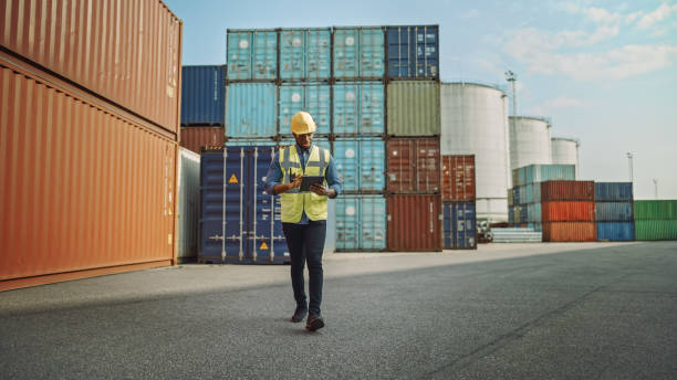 przystojny african american black industrial engineer w żółty hard hat i kamizelka bezpieczeństwa pracy na tablecie. foreman lub inspektor chodzenie w pracy w terminalu kontenerowym. - commercial dock audio zdjęcia i obrazy z banku zdjęć