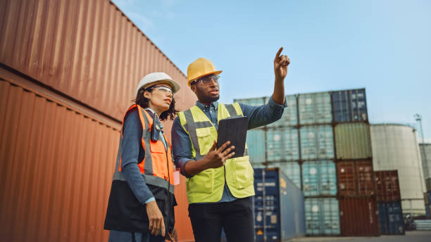 Multiethnic Female Industrial Engineer with Tablet and Black African American Male Supervisor in Hard Hats and Safety Vests Stand in Container Terminal. Colleagues Talk About Logistics Operations. Multiethnic Female Industrial Engineer with Tablet and Black African American Male Supervisor in Hard Hats and Safety Vests Stand in Container Terminal. Colleagues Talk About Logistics Operations. harbor stock pictures, royalty-free photos & images