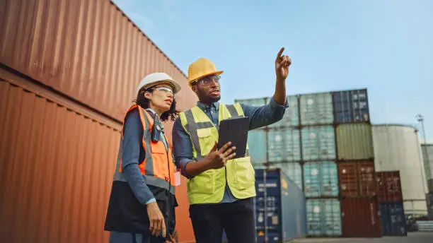 Photo of Multiethnic Female Industrial Engineer with Tablet and Black African American Male Supervisor in Hard Hats and Safety Vests Stand in Container Terminal. Colleagues Talk About Logistics Operations.