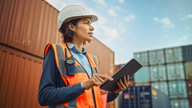 glimlachend portret van een mooie latijnse vrouwelijke industriële ingenieur in witte harde hoed, high-visibility vest dat op tabletcomputer werkt. inspecteur of veiligheidstoezichthouder in containerterminal. - vrachtcontainer stockfoto's en -beelden