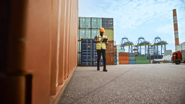 black african american mężczyzna industrial supervisor w hard hat i kamizelki bezpieczeństwa jest za pomocą tabletu podczas spaceru obok kontenera w logistics operations terminal. - commercial dock audio zdjęcia i obrazy z banku zdjęć