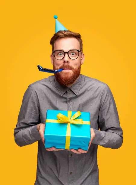 Excited young bearded male in casual shirt and eyeglasses with party hat and noisemaker holding gift box and looking at camera on yellow background