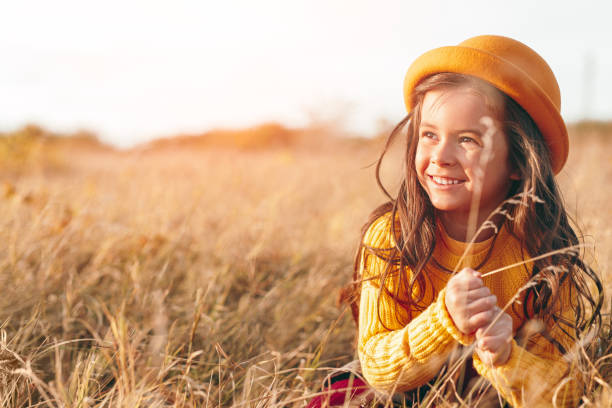 Positive kid in trendy hat smiling and looking away while playing with dry grass in summer evening in meadow