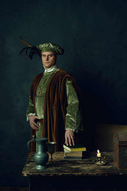 istock Renaissance man stands behind a table with books on it, a tin pitcher, a tin goblet, a candlestick and an iron box. 1309571679