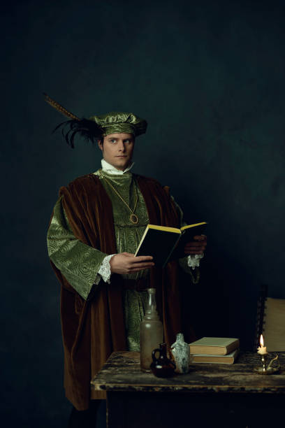 istock Renaissance man with a book in his hands stands behind a table with books on it, three glass bottles, and a candlestick. 1309571682