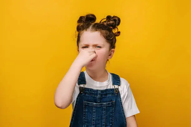 Photo of Unhappy sad little preschool girl kid feeling bad and dislike smell and hold nose from funny odor, dressed in white t-shirt, isolated on yellow studio background, six year child looking at camera