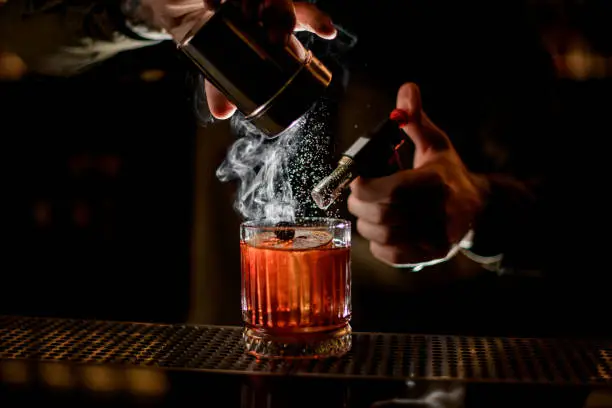 Photo of bartender carefully sprinkles icing sugar on steaming glass with alcoholic cocktail on the bar counter