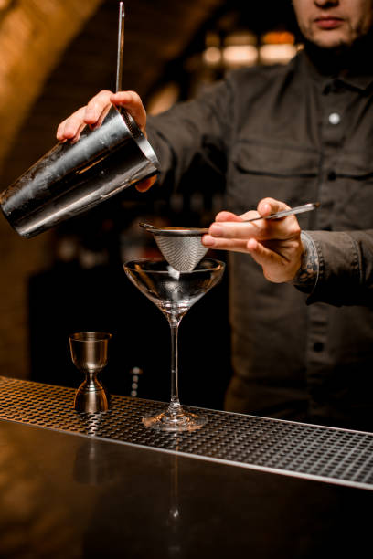 man bartender holds sieve and shaker cup over martini glass - transparent holding glass focus on foreground imagens e fotografias de stock