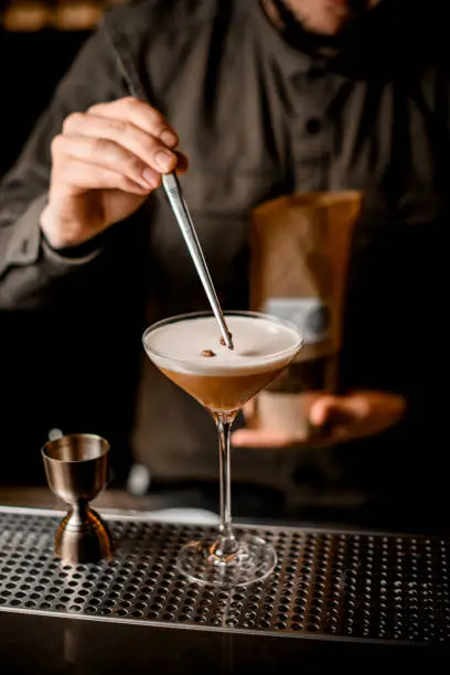 view on glass with frothy espresso martini cocktail which bartender neatly decorating with coffee bean using tweezers