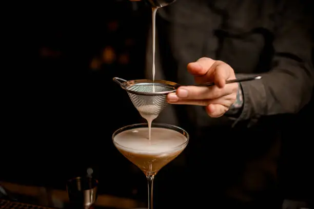 close-up view on metal sieve through which male bartender pours frothy espresso martini cocktail into glass