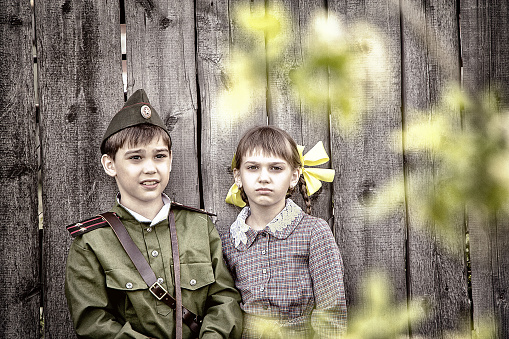 Postcard, stylized as vintage for the Victory Day. A boy in a military uniform and a girl in an old dress are standing near an old fence. The theme of May 9, Victory Day in Russia. Soft selective focus, added noise.