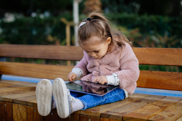 Adorable little girl in a dress sitting under a small tree, leaning on a tree trunk and looking down while holding her smart phone and texting in a beautiful sunset in summertime.