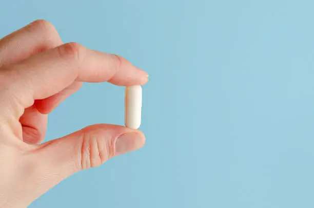 Female hand holding white capsule on blue background. Close, copy space. Medicine concept