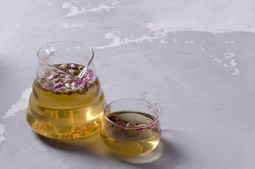 A glass kettle and a small glass cup with brewed tea made of rose petals stand on a gray concrete background with a copy of the space, selective focus