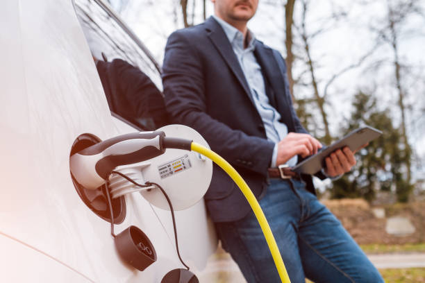 Business man standing near charging electric car or EV car and using tablet in the street at the sunlight Business man standing near charging electric car or EV car and using tablet in the street at the sunlight. ev charging stock pictures, royalty-free photos & images