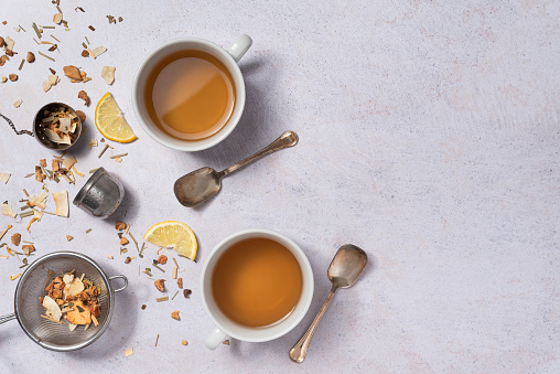 Two cups of tea, slices of lemon, spoon with infuser and strainer on a light gray table, flat lay.