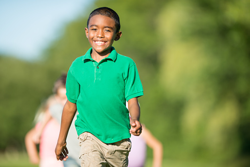 An adorable four year old boy is at the park with his friends on a summer day. He is dressed casually and smiling at the camera.