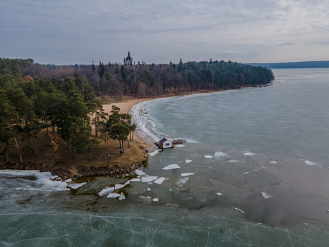 Aerial view of cracked ice on Kaunas lake and coastline with a hut and a footbridge from above