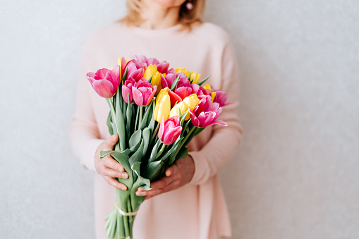 Portrait of cropped woman with blonde hair hiding her face, and holding bouquet of pink and yellow tulip flowers. White background, copy space, close up.