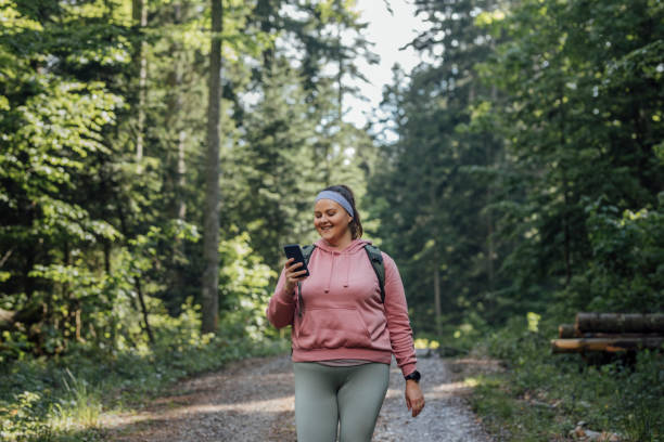 mode de vie sain et actif : une belle femme de taille heureuse plus dans un sweat-shirt rose marchant dans la forêt et utilisant un téléphone portable - spring women relaxation people photos et images de collection