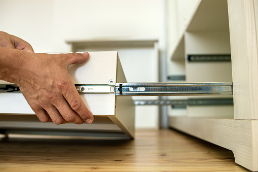 Close up of carpenter hands installing wooden drawer on sliding skids in contemporary cupboard cabinet.