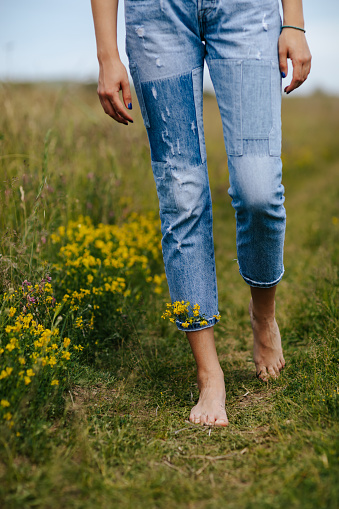 Carefree young woman spinning in idyllic rural field of wildflowers. Close-up young female legs walking on green spring grass with wildflowers in legs of denim trousers