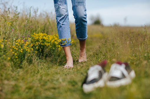 Carefree young woman spinning in idyllic rural field of wildflowers. Close-up young female legs walking on green spring grass with wildflowers in legs of denim trousers