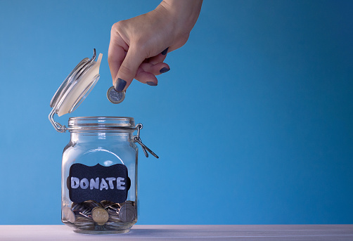 Hand putting a coin in a glass jar with coins with a chalk donate tag on a blue background, donation and charity concept. Copy space.