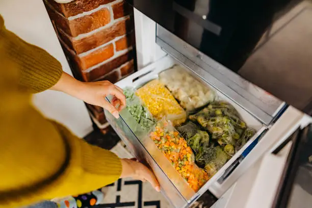 Woman putting container with frozen mixed vegetables to refrigerator