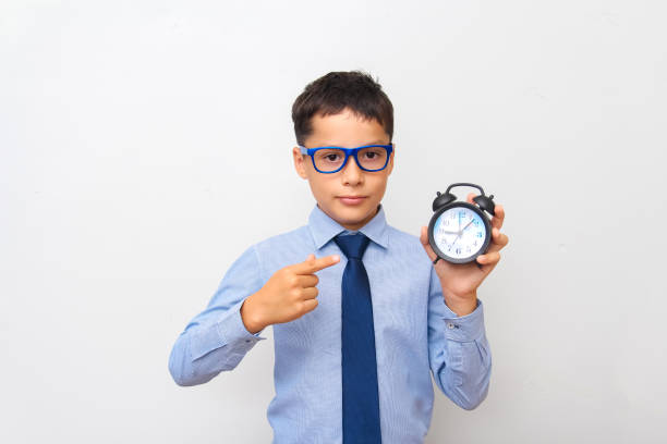 파란 셔츠와 안경을 쓴 검은 머리의 백인 소년이 손에 알람 시계를 들고 회색 배경을 손가락으로 가리킨다. 마감일, 시간 관리의 개념 - focus on foreground alarm clock deadline time 뉴스 사진 이미지