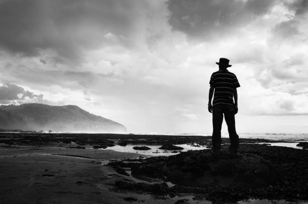 czarno-biały wizerunek mężczyzny stojącego na skałach i obserwującego zbliżającą się burzę na plaży muriwai, west auckland - murawai beach zdjęcia i obrazy z banku zdjęć