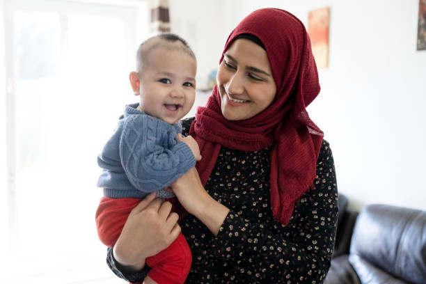 Candid portrait of British Asian mother and baby son at home Partial front view of early 30s woman in hijab and tunic smiling at 5 month old boy in her arms as he looks at camera and laughs. pakistani ethnicity stock pictures, royalty-free photos & images