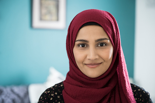 Close-up of early 30s woman wearing maroon headscarf and tunic standing in natural light of family home looking at camera with contented smile.