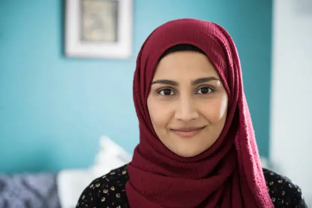 Close-up of early 30s woman wearing maroon headscarf and tunic standing in natural light of family home looking at camera with contented smile.