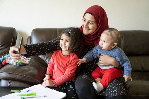Partial front view of mid adult woman in headscarf and tunic sitting on living room sofa with baby son and toddler daughter smiling at smartphone camera.
