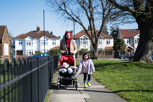Full length front view of early 30s mother, 2 year old daughter, and 5 month old son in stroller approaching camera on late winter walk in West London suburb.