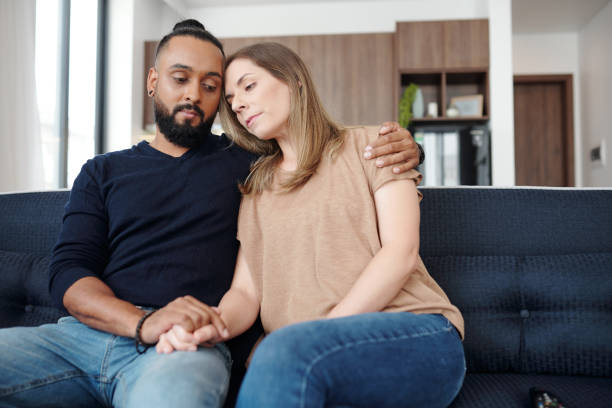Couple suffering from loss Sad young mixed-race couple reassuring each other when sitting on sofa at home miscarriage stock pictures, royalty-free photos & images