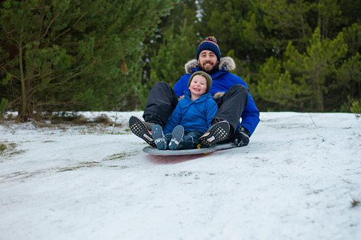 An uncle and nephew wearing warm winter clothing in a snowy, rural, outdoor setting. They are having fun sledding down a snowy hill.