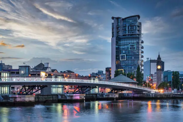 Belfast City Sunset with colorful twilight over Lagan Weir Pedestrian and Cycle Bridge spanning over the Lagan River in downtown Belfast. Illuminated Modern Urban Riverside Cityscape of Belfast. Belfast, Northern Ireland, UK, Europe