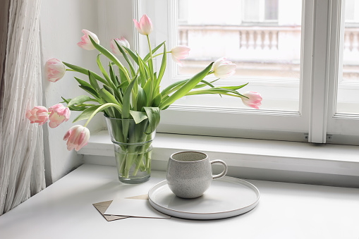 Easter spring still life. cup of coffee and floral bouquet on window sill. Pink tulips flowers in ceramic vase pot. Blank greeting card mockup, home decor, breakfast concept. Scandinavian interior